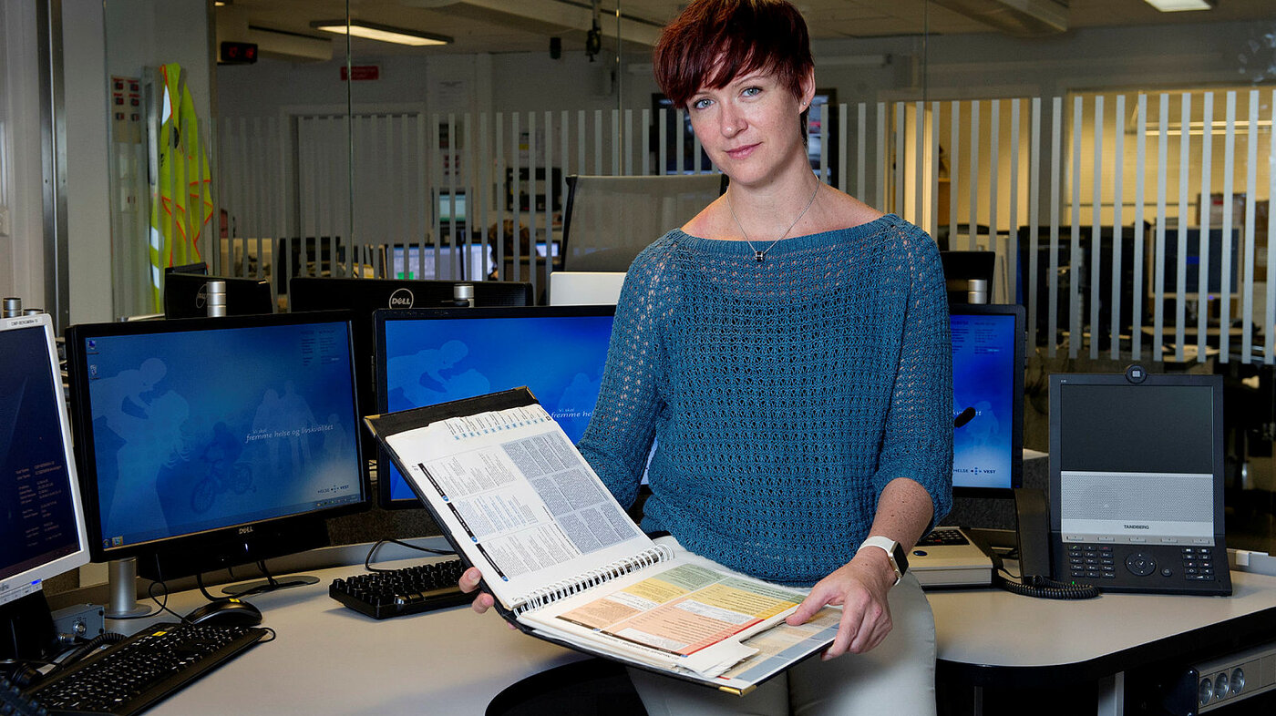 Bildet er et portrett av lege Eirin Ellensen på en AMK-sentral med Norsk indeks for medisinsk nødhjelp i hendene.