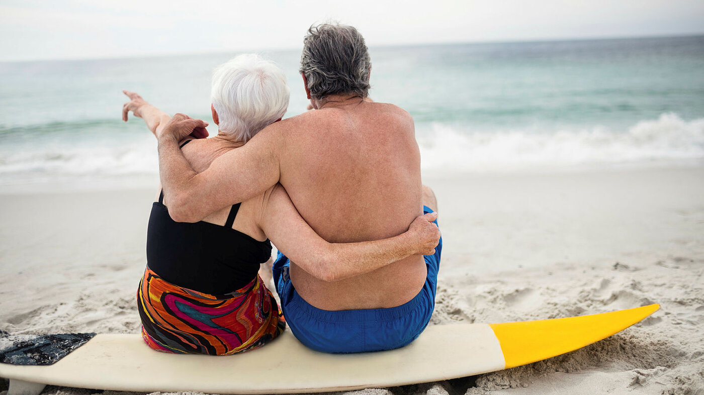 Bildet viser et eldre par som sitter på et surfebrett og ser ut over havet og bølgene