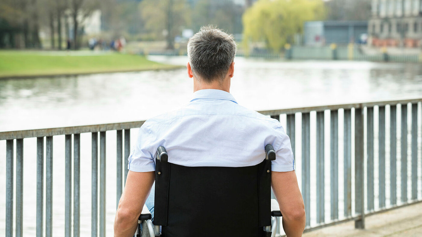 Bildet viser en mann sett bakfra som sitter i en rullestol og ser ut over en elv.