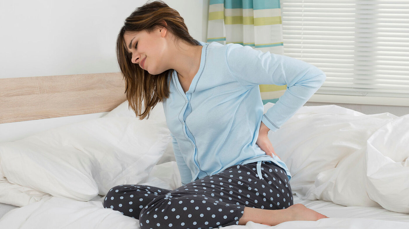 Bildet viser en ung kvinne som sitter i sengen og har vondt i ryggen.