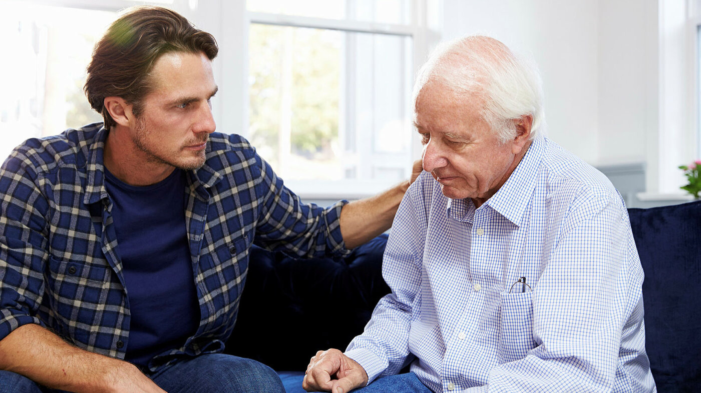 Bildet viser en sønn som snakker med en eldre, syk og nedfor far. Han holder armen trøstende på skulderen til faren.