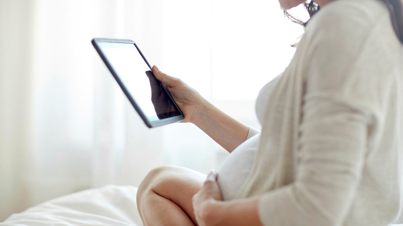Bildet viser en gravid kvinne som sitter i en seng og ser på et nettbrett