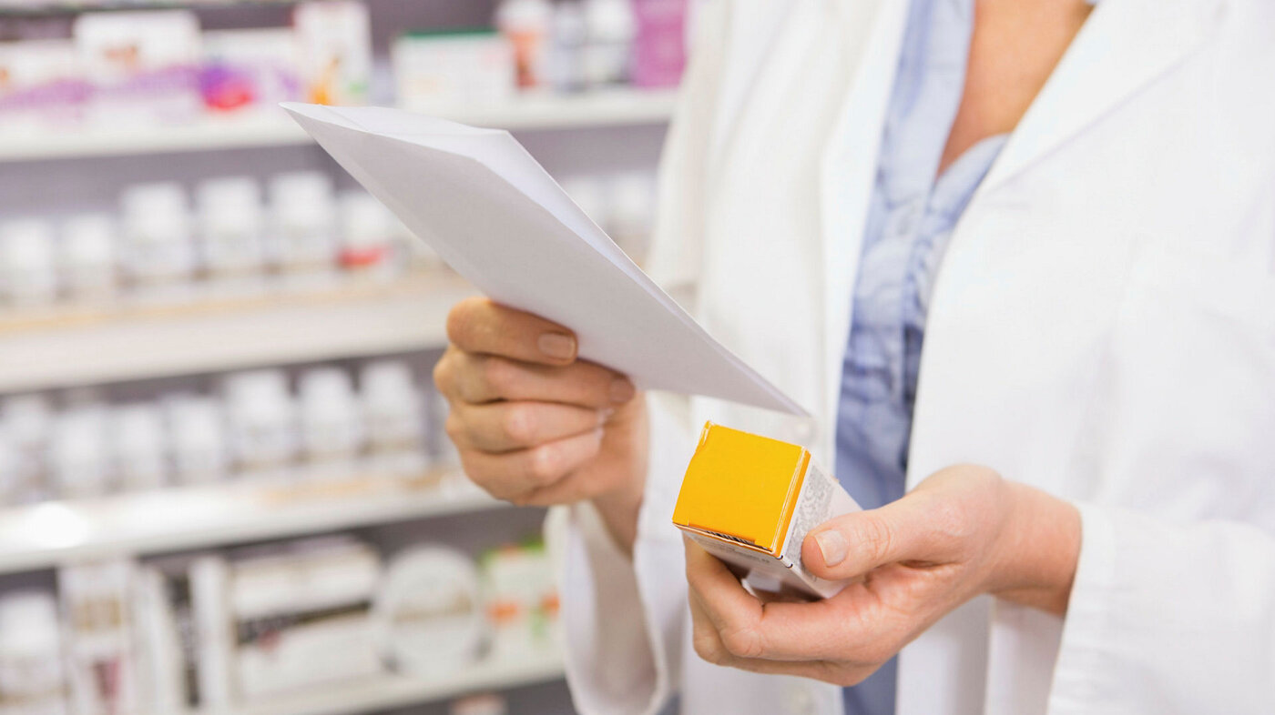 Bildet viser hendene til en farmasøyt som holder på en eske tabletter og leser på et ark. Hun er på et apotek.