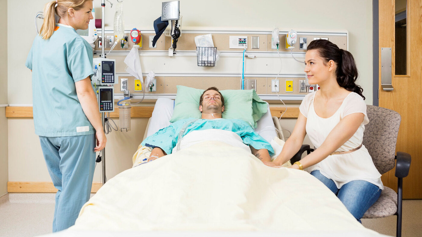 Bildet viser en pasient i sengen, mens en pårørende holder ham i hånda. Ved siden av står en sykepleier.