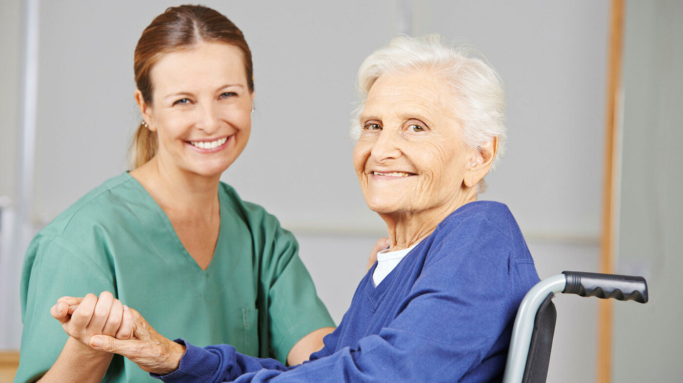Bildet viser en sykepleier som holder en eldre pasient i hånda. Pasienten sitter i rullestol, og begge smiler.