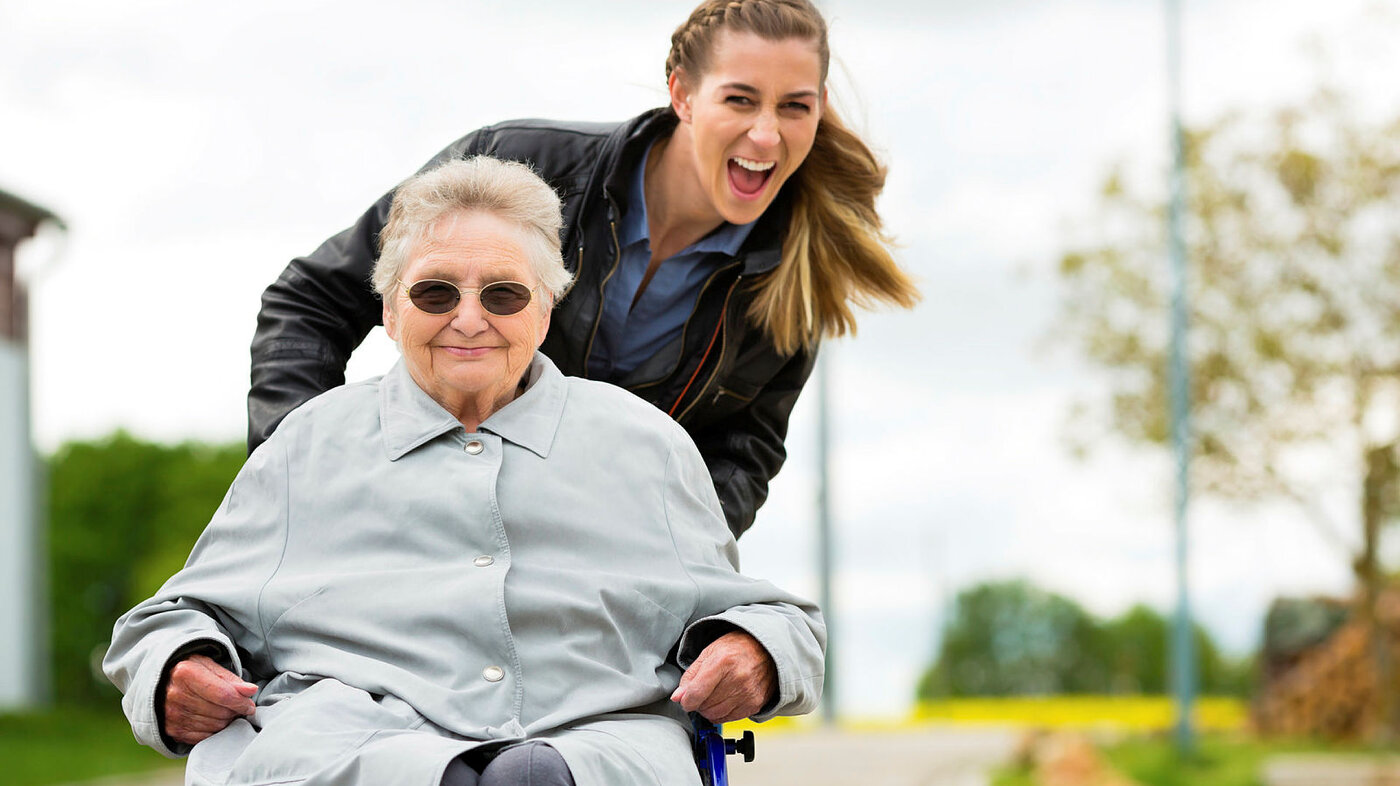 Bildet viser en eldre dame i rullestol som er ute på tur med en ung kvinne.