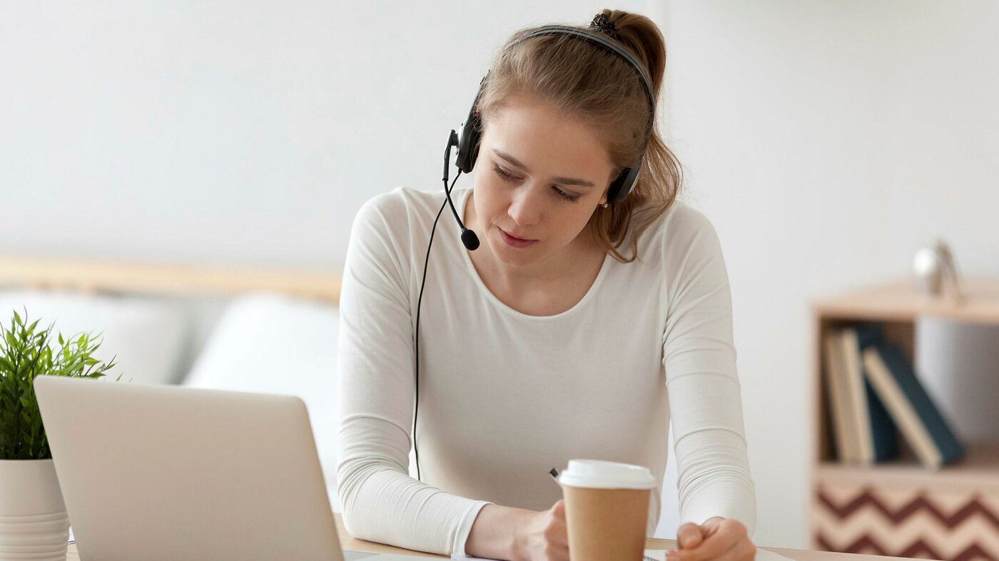 Bildet viser en ung kvinne som sitter i stua foran PC-en. Hun har hodetelefon med mikrofon på.