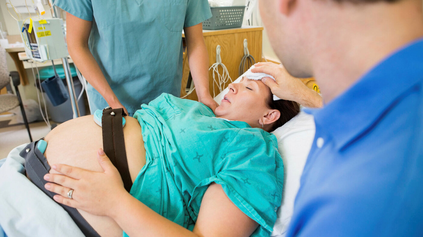 Bildet viser en kvinne som holder på å føde. Sykepleierne og legen måler fosterlyden, mens faren tørker pannen til kvinnen.