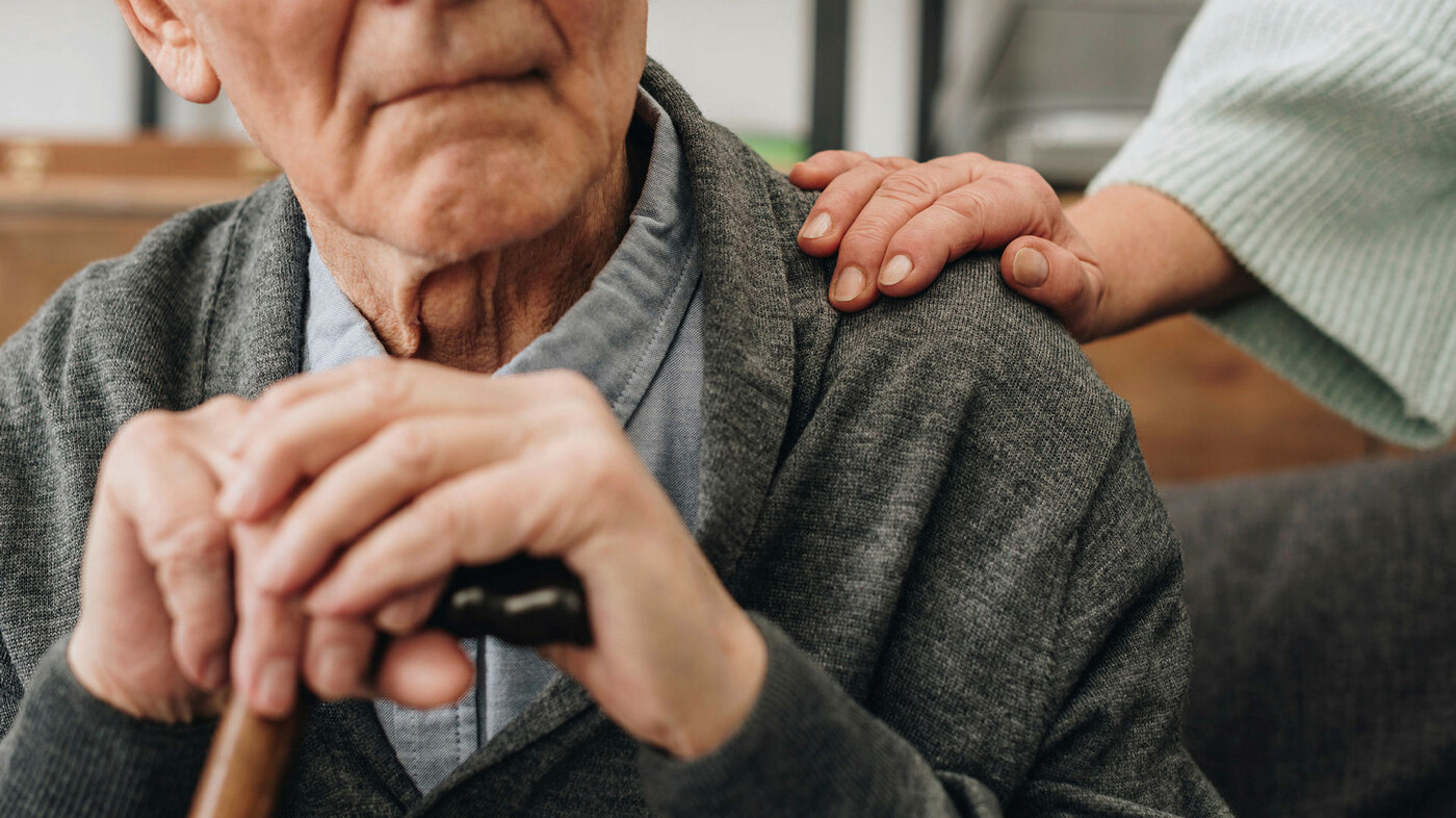Bilder viser et utsnitt av ansiktet til en eldre mann. Han holder hendene over håndtaket på stokken sin. En trøstende hånd ligger på skulderen.