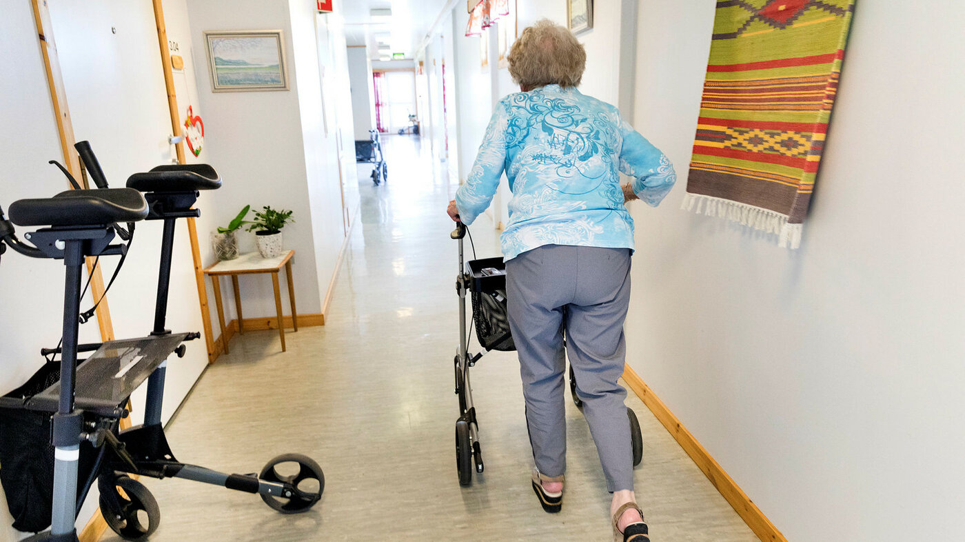 Eldre kvinne med gåstol i korridor fotografert bakfra.