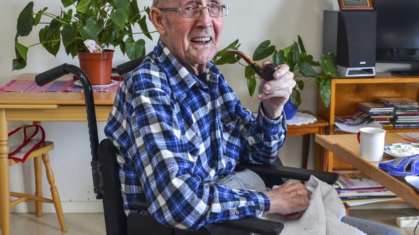 John Gundersen i omsorgsboligen sin Bildet viser John Gundersen på 93 år. Han sitter i en rullestol i omsorgsboligen sin. Han har en pipe i hånden og smiler til fotografen.