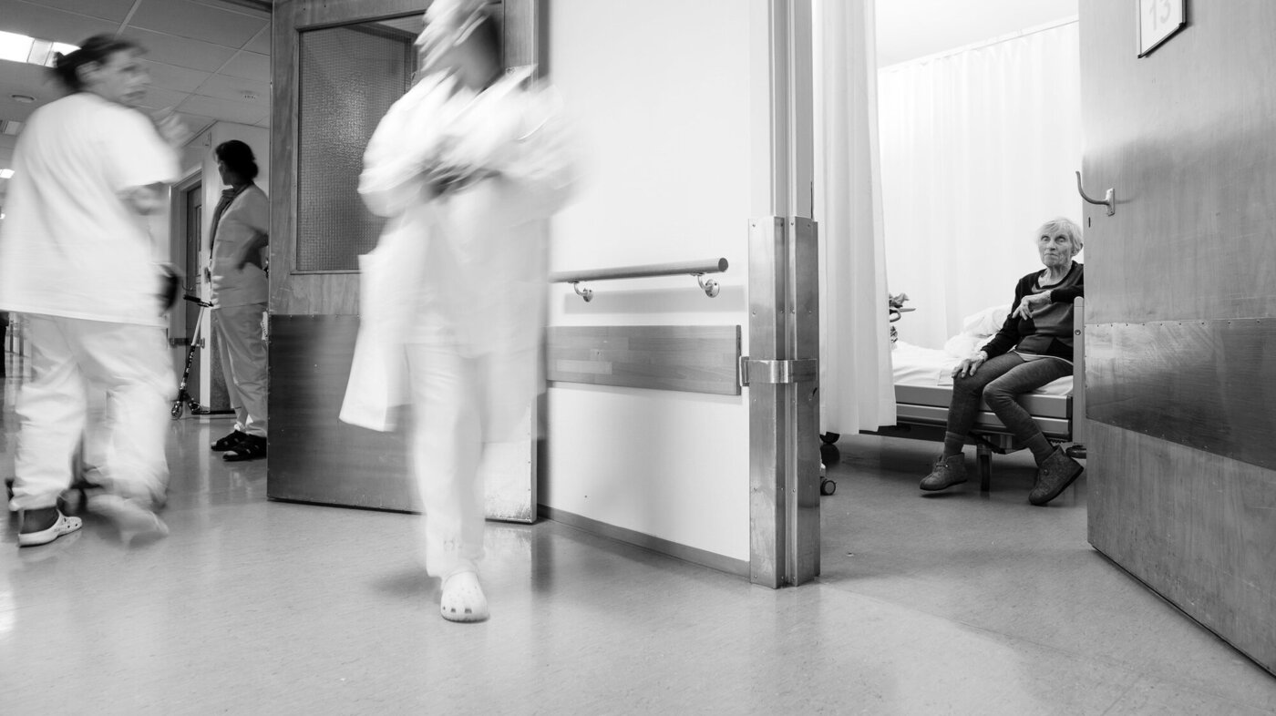 Bildet viser pasient i senga som venter på hjelp fra opptatt personale som fyker forbi.