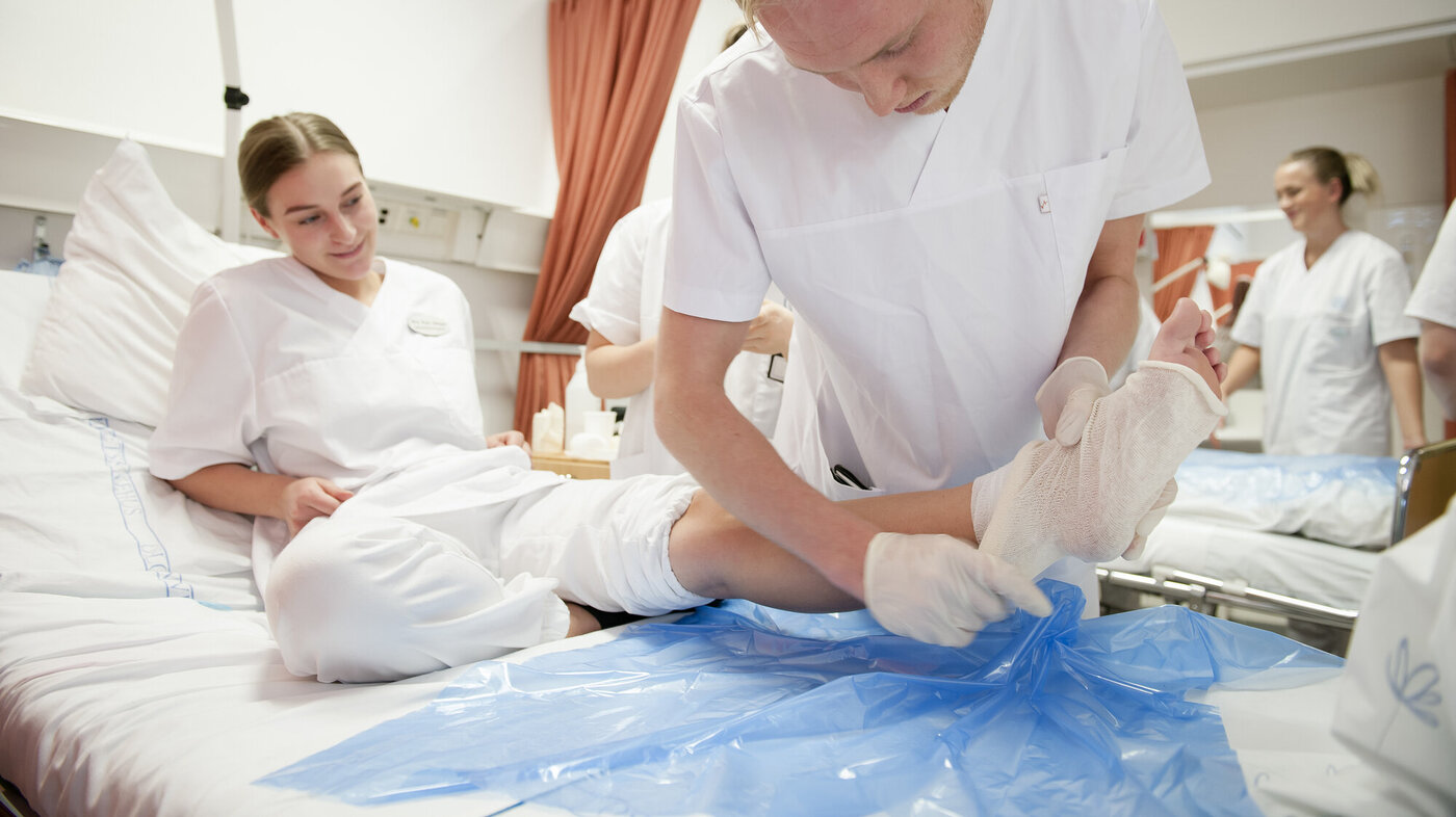 Sykepleierstudenter i praksis. Kvinne ligger med bandasje i sykeseng, mannlig student jobber konsentrert med en bandasje rundt foten. 