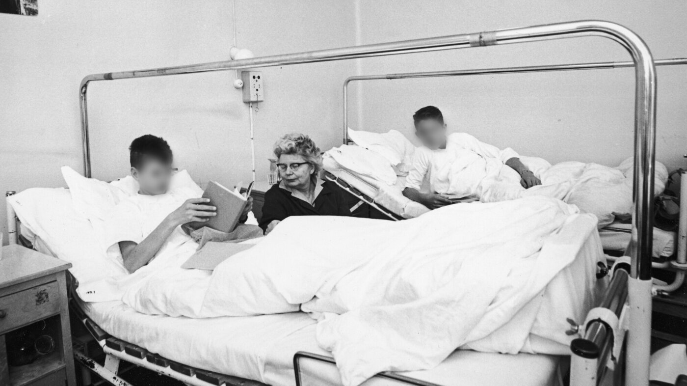 Bildet viser to gutter i hver sin sykehusseng, en eldre kvinne sitter mellom dem.
