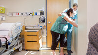 Bildet viser en høygravid kvinne som står ved vinduet. En sykepleier holder armen beskyttende rundt henne.