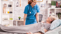 Bildet viser en sykepleier eller annet helsepersonell som står over sengen til en eldre dame på sykehjem eller hjemme hos seg selv