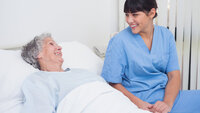 Bildet viser en eldre kvinne som ligger i en seng og smiler til et yngre kvinnelig helsepersonell som smiler tilbake