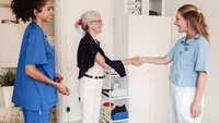 Bildet viser en eldre kvinne som tar et helsepersonell i hånden mens at annet helsepersonell ser på