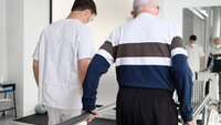 Bildet viser et helsepersonell som hjelper en eldre mann med å øve på å gå 