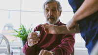 Bildet viser en eldre mann som får et glass med medisiner av helsepersonell