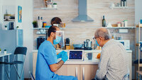 Bildet viser en mannlig sykepleier i blå uniform, munnbind og visir på hjemmebesøk hos en eldre mann, også han med munnbind. De sitter og ser på en ipad.