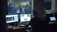 Bildet viser en operatør av en simulering, som sitter og styrer det hele via tre dataskjermer, foran et ensidig speil. På andre siden foregår simuleringen