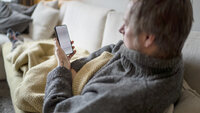 Bildet viser en mann som ligger under et pledd på sofaen og ser på mobilen