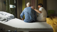 Bildet viser en sykepleier som sitter på sengekanten sammen med en eldre pasient