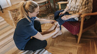 Bildet viser en hjemmesykepleier som skifter strømper på en eldre dame som sitter i stolen i hjemmet sitt.