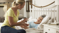 Bildet viser en eldre, syk dame i senga. På sengekanten sitter dattera