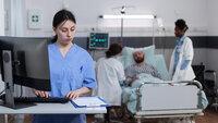 Bildet viser en sykepleier som står på et pasientrom foran en PC. Foran henne ligger et skjema. Bak henne ligger en pasient i senga med to sykepleiere ved sengekanten