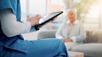 Bildet viser en sykepleier som sitter med en iPad med en pasient i bakgrunnen
