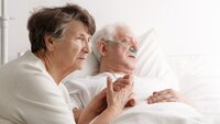 Bildet viser en kone som sitter ved sykesengen til sin døende mann