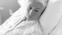 Bildet viser nærbilde av ei eldre dame i sengen, som virker ettertenksom