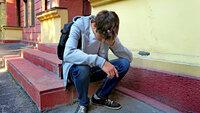 Bildet viser en tenåringsgutt med ransel som sitter på en trapp utenfor skolen. Han er lei seg.