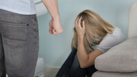 Bildet viser en mann som står med knyttneven foran en redd dame som har krøket seg sammen bak sofaen.