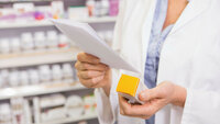 Bildet viser hendene til en farmasøyt som holder på en eske tabletter og leser på et ark. Hun er på et apotek.