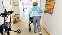 Eldre kvinne med gåstol i korridor fotografert bakfra.