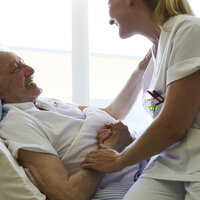 Bildet viser en pasient som ligger i senga, og sykepleieren sitter på sengekanten og holder ham på armen. Begge ler.