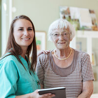 En eldre kvinne og en ung, kvinnelig sykepleier smiler og ser inn i kamera.