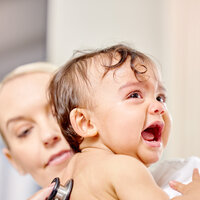 Bildet viser et lite barn som gråter mens en sykepleier lytter på ryggen til barnet