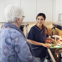 Bildet viser en kvinnelig sykepleier som smiler og snakker med en eldre kvinne