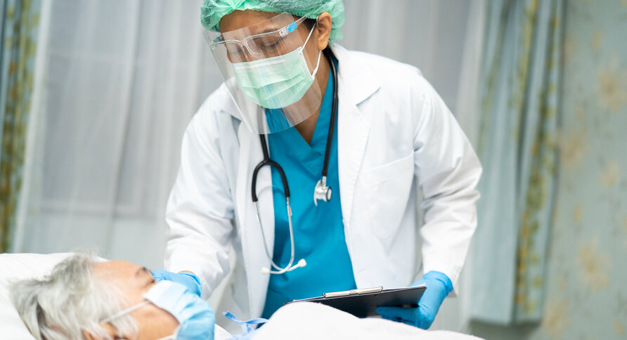Bildet viser en eldre mann som ligger i senga med maske på, mens legen står ved siden av med maske og visir