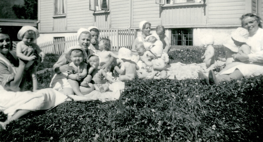 Historisk, gammelt bilde av sykepleiere med små barn på gresset utenfor Bergen kommunale mødrehjem.